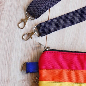 May include: A rainbow striped zippered pouch with a blue strap and two silver clasps. The pouch is attached to two black straps with silver clasps.