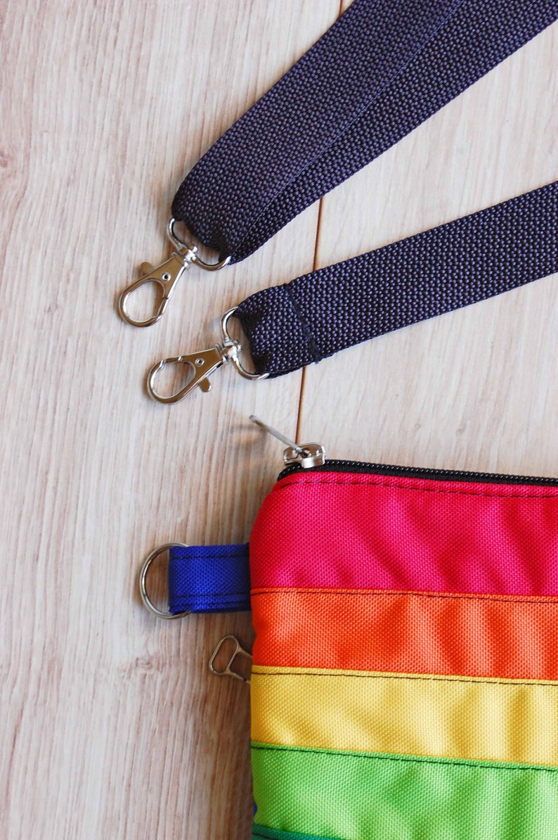 May include: A rainbow striped zippered pouch with a blue strap and two silver clasps. The pouch is attached to two black straps with silver clasps.