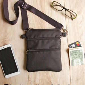 May include: A black crossbody bag with three zippered compartments. The bag has a long adjustable strap and is shown on a wooden surface with a cell phone, glasses, and cash.
