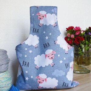 Pode incluir: Uma bolsa de projeto de tecido azul com um padr&atilde;o de ovelhas brancas e nuvens. A bolsa tem uma al&ccedil;a e diz "BAA" nela. A bolsa est&aacute; sentada em uma superf&iacute;cie de madeira com fio e agulhas de tric&ocirc;.
