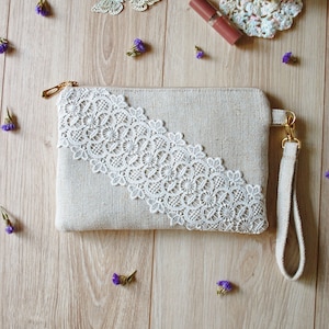 May include: A beige fabric clutch with a white lace trim and a long beige strap. The clutch is on a wooden surface with small purple flowers.