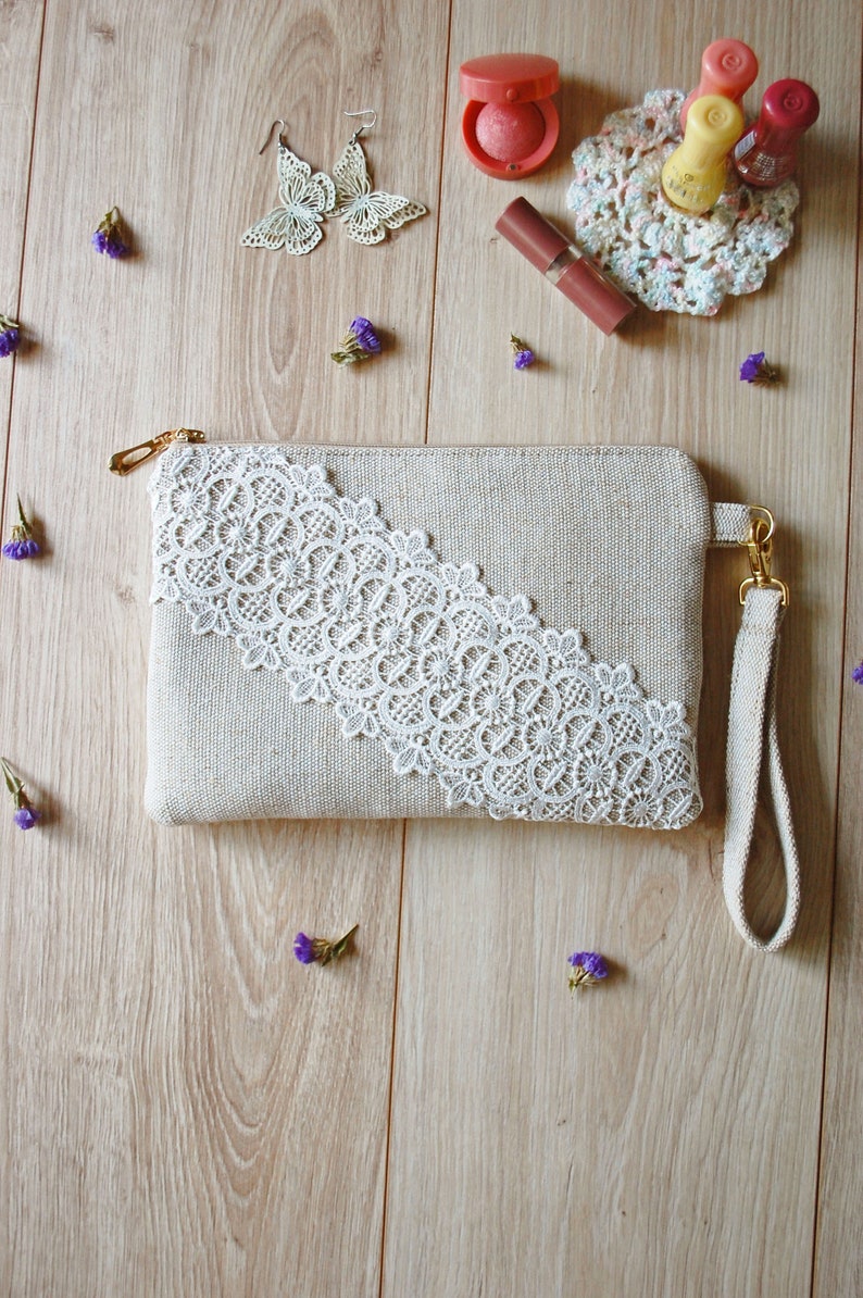 May include: A beige fabric clutch with a white lace trim and a long beige strap. The clutch is on a wooden surface with small purple flowers.