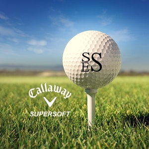 May include: A white Callaway Supersoft golf ball with the letters "SS" and "E" printed on it. The ball is on a tee and sitting on a green grassy surface.
