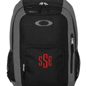 May include: A black and gray backpack with a large gray Oakley logo on the front. The backpack has a zippered pocket on the front and a mesh pocket on the side. The backpack has a red monogram "SSE" on the front.