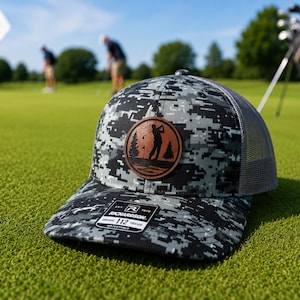 May include: A gray and black digital camouflage trucker hat with a brown circular leather patch featuring a silhouette of a golfer, trees, and a flag. The hat has a mesh back and a black sticker on the brim with the text Richardson 112.