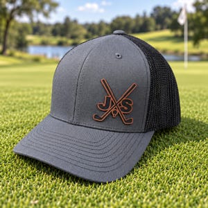May include: A charcoal gray baseball cap with a black mesh back and a brown leather patch on the front. The patch features a graphic of two crossed golf clubs with a golf ball in the center and the letters J and S positioned on either side.