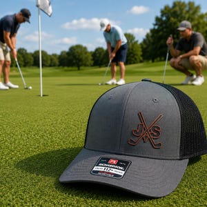 Peut inclure: Une casquette trucker gris anthracite et noir avec un écusson en cuir marron portant les lettres JS et deux clubs de golf croisés. La casquette présente un panneau avant structuré, une visière courbée et un filet noir avec une étiquette Richardson 112.