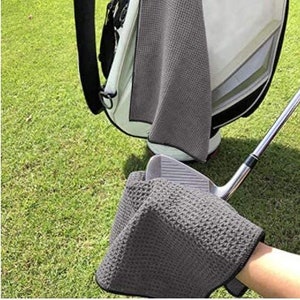 Puede incluir: Una toalla de golf de microfibra gris con textura de gofre. La toalla est&aacute; doblada y colocada encima de un palo de golf.
