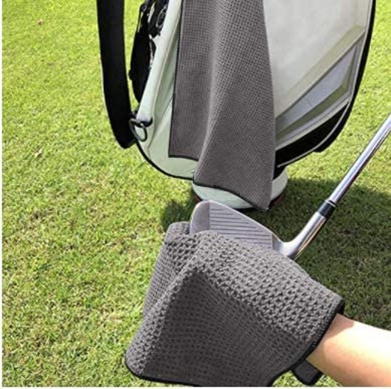Puede incluir: Una toalla de golf de microfibra gris con textura de gofre. La toalla est&aacute; doblada y colocada encima de un palo de golf.
