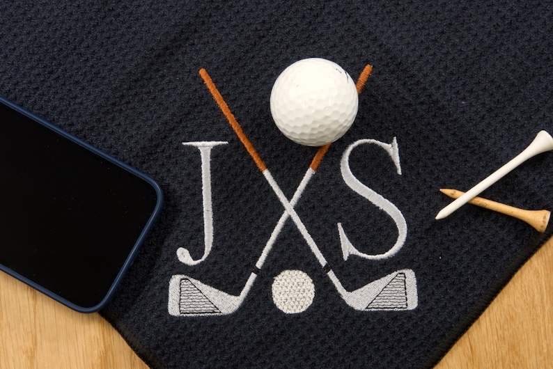 Puede incluir: Una toalla de golf negra con palos de golf bordados en blanco, una pelota de golf y las letras JXS. Dos tees de golf est&aacute;n en la toalla.