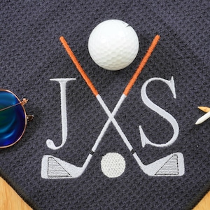 Puede incluir: Una toalla de golf gris oscuro con las iniciales "JXS" bordadas en blanco. Dos palos de golf se cruzan detr&aacute;s de las iniciales, y una bola de golf blanca est&aacute; centrada entre los palos. Un tee de golf blanco y uno marr&oacute;n est&aacute;n en la esquina inferior derecha de la toalla.