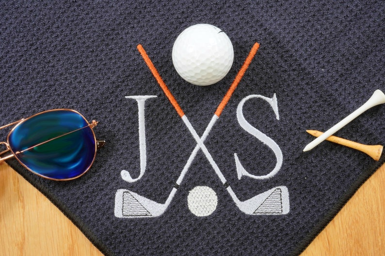 Puede incluir: Una toalla de golf gris oscuro con las iniciales "JXS" bordadas en blanco. Dos palos de golf se cruzan detr&aacute;s de las iniciales, y una bola de golf blanca est&aacute; centrada entre los palos. Un tee de golf blanco y uno marr&oacute;n est&aacute;n en la esquina inferior derecha de la toalla.