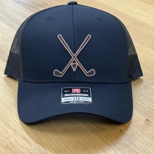 May include: A navy blue trucker hat with a mesh back and a curved brim. The front features a brown golf club and ball design. The hat has the text "RICHARDSON" and "ORIGINAL 112 TRUCKER ADJUSTABLE".