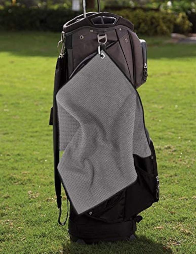 Puede incluir: Una toalla de golf de microfibra gris con borde negro cuelga de una bolsa de golf. La toalla est&aacute; unida a un mosquet&oacute;n plateado.
