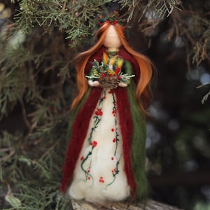 May include: A needle-felted doll with long orange hair and a floral crown, wearing a cream-colored gown with red berry and green vine embroidery, layered under a dark green and deep red cloak, holding a small basket of colorful dried flowers.