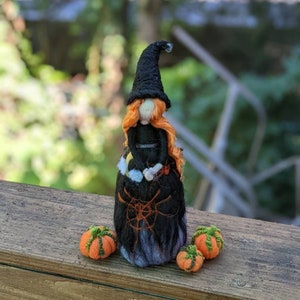 May include: A small felt witch doll with orange hair, a black hat, and a black dress with a spiderweb design. The doll is sitting on a wooden surface with three small orange pumpkins.