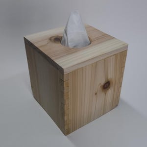 May include: A square wooden tissue box with a natural wood finish. The box has a rectangular opening on top with a white tissue protruding. The wood grain and knots are visible, adding to the rustic aesthetic.