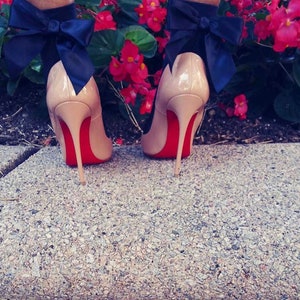 Peut inclure: Une paire de chaussures &agrave; talons hauts couleur nude avec des semelles rouges et un grand n&oelig;ud noir &agrave; l'arri&egrave;re de chaque chaussure.