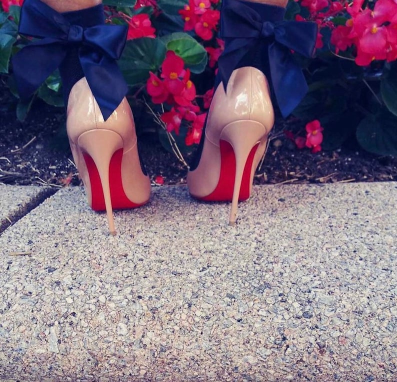 Peut inclure: Une paire de chaussures &agrave; talons hauts couleur nude avec des semelles rouges et un grand n&oelig;ud noir &agrave; l'arri&egrave;re de chaque chaussure.