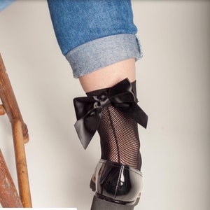 Peut inclure: Chaussettes noires en r&eacute;sille avec un grand n&oelig;ud en satin noir &agrave; la cheville. Les chaussettes sont port&eacute;es avec des chaussures en cuir verni noir.