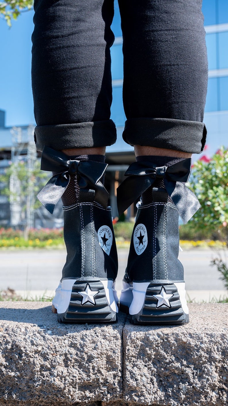 Peut inclure: Des baskets Converse noires montantes avec des &eacute;toiles blanches et un ruban noir nou&eacute; en n&oelig;ud autour de la cheville.