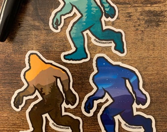 May include: Three vinyl stickers featuring a Bigfoot silhouette with different landscape backgrounds. The stickers are in a blue, brown, and purple color scheme. The text "KNOWSQUATCH" is printed on each sticker.