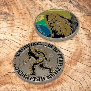 May include: Two silver-colored challenge coins with a Sasquatch theme. One coin features a colorful illustration of a Sasquatch with the text "Inknowatch" and "Sasquatch" on the side. The other coin features a black and gold illustration of a Sasquatch walking with the text "Seeing is believing... is better than believing" and "Est. 2014".