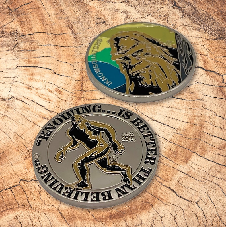 May include: Two silver-colored challenge coins with a Sasquatch theme. One coin features a colorful illustration of a Sasquatch with the text "Inknowatch" and "Sasquatch" on the side. The other coin features a black and gold illustration of a Sasquatch walking with the text "Seeing is believing... is better than believing" and "Est. 2014".