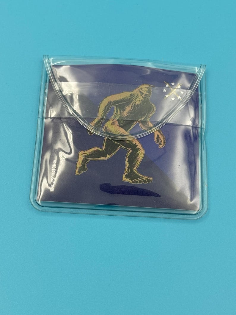 May include: A clear plastic pouch with a blue background and a graphic of a Bigfoot walking. The graphic is in gold and brown colors. The text "IN OUSSMATCHING" is visible on the pouch.