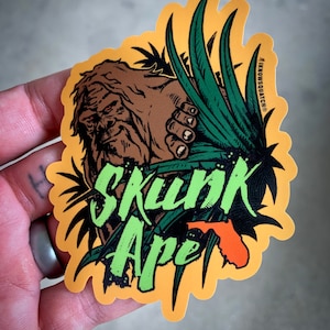 May include: A yellow sticker with a green and brown illustration of a creature with a hand reaching out from behind green leaves. The text "Skunk Ape" is written in green with a stylized orange outline of the state of Florida.