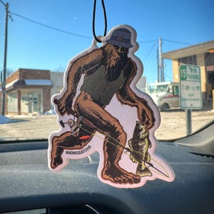 May include: Air freshener featuring a cartoon Bigfoot figure fishing. The brown and tan figure wears a hat and vest, holding a fishing rod with a caught fish. The text "IKNOWSQUATCH" is printed on the bottom.