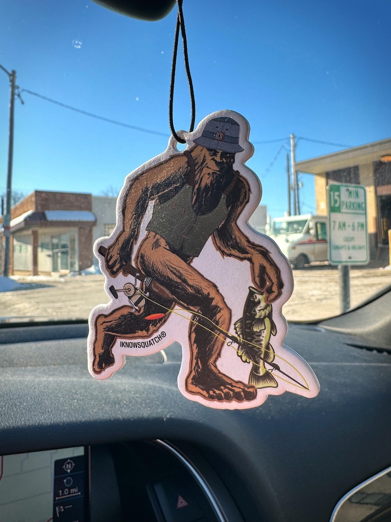 May include: Air freshener featuring a cartoon Bigfoot figure fishing. The brown and tan figure wears a hat and vest, holding a fishing rod with a caught fish. The text "IKNOWSQUATCH" is printed on the bottom.