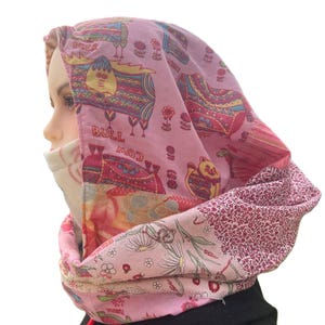 May include: A pink and patterned hood scarf with a drawstring. The scarf features a patchwork design with cow and floral motifs. The hood is lined with a cream-colored fabric. The scarf is worn over a black garment.