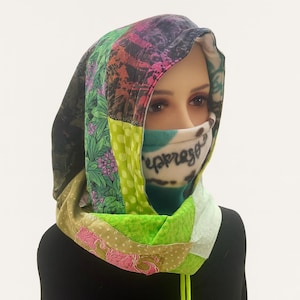 May include: A patchwork hood with a face covering. The hood features a variety of fabrics in shades of green, pink, and purple. The face covering has the word "espresso" printed on it. A yellow drawstring is visible.
