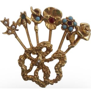 Può includere: Una spilla color oro con una base a nastro testurizzata che sostiene cinque steli verticali. Gli steli sono sormontati da un rametto con perle, un'ape con una pietra verde, un serpente con una pietra blu, un fiore rosso e un fiore blu.
