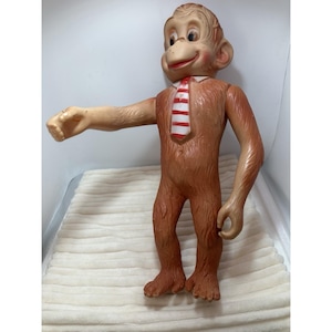 May include: A brown monkey figurine with a molded fur texture, featuring a smiling face with pink cheeks and a white tie with horizontal red stripes. The monkey is standing upright with one arm extended to the side and the other arm at its side.