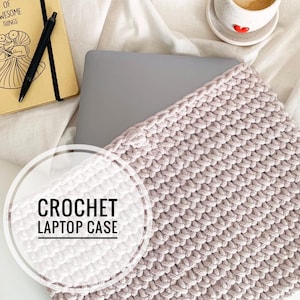 Può includere: Una custodia per laptop in crochet di colore rosa chiaro. La custodia è realizzata con un semplice motivo a punto e ha un aspetto testurizzato. La custodia è appoggiata su una superficie bianca con un laptop e un quaderno visibili sullo sfondo.