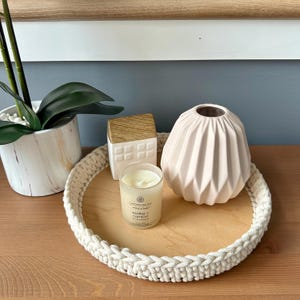 以下が含まれることがあります： 木製の土台に白い編み込みのトレーがあり、白いろうそく、ピンクの陶器の花瓶、木製の屋根が付いた白い陶器の家が入っています。