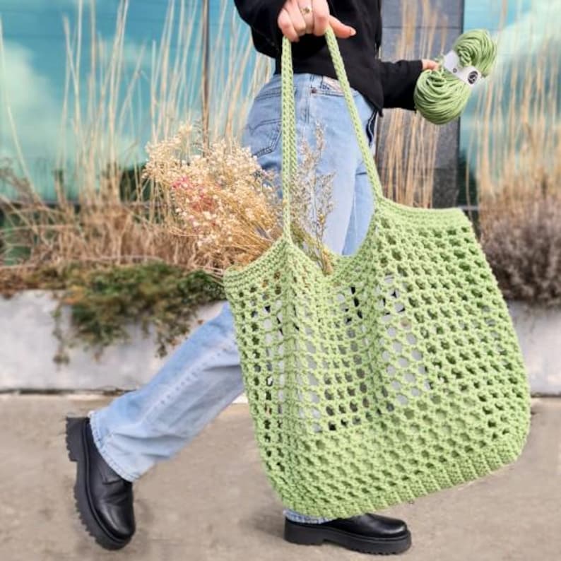 May include: A light green crochet tote bag with a large open weave pattern. The bag is being held by a person wearing blue jeans.