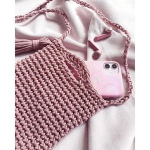 May include: A pink crocheted bag with a tassel and a phone case inside. The bag has a long strap and is made of a soft, textured yarn.