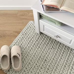 May include: A light gray, chunky knit rug with a textured surface. The rug is placed on a light wood floor and has two pairs of beige slippers on top of it.