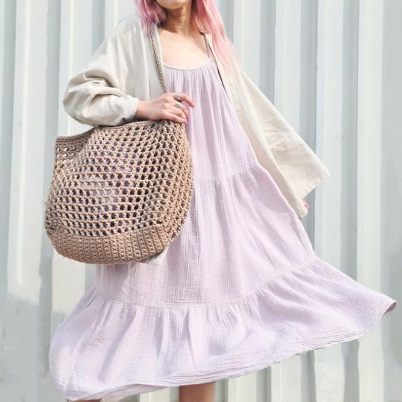 May include: A woman wearing a light purple tiered dress and a beige cardigan holds a large brown crocheted tote bag.