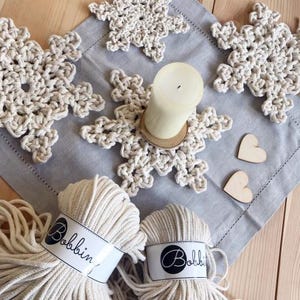 May include: Four white crocheted snowflake coasters with a white candle in the center. Two balls of white cotton rope labeled "Bobbin'" are in the bottom of the image.