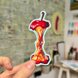 May include: A sticker showing a colourful illustration of an apple core. The artwork depicts the core in shades of red, orange, and brown, with a light blue outline. The sticker is held by a hand against a blurred background.
