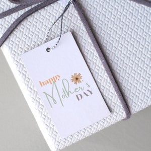 May include: A white gift tag with a gray ribbon attached. The tag has the text "happy Mother's Day" in green and orange lettering with a small flower graphic.