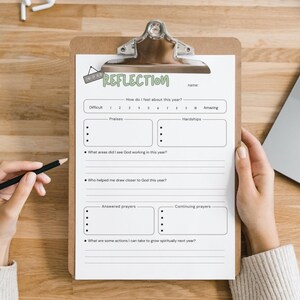 May include: A clipboard with a white paper titled "REFLECTION" is held by a person. The paper has sections for "Praises," "Hardships," and prompts about spiritual growth. A pencil rests on the wooden surface, near the paper. A laptop is visible in the background.