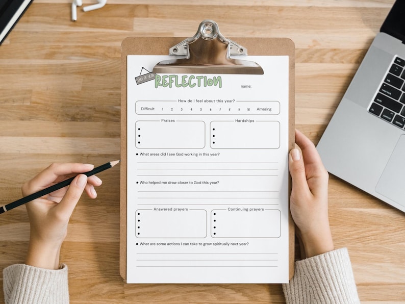 May include: A clipboard with a white paper titled "REFLECTION" is held by a person. The paper has sections for "Praises," "Hardships," and prompts about spiritual growth. A pencil rests on the wooden surface, near the paper. A laptop is visible in the background.