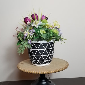 May include: A black and white geometric planter with a floral arrangement of purple, green, and white flowers sits on a wooden cake stand with a black pedestal base.