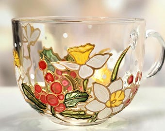 May include: A clear glass teacup with a handle, adorned with hand-painted floral designs. The cup features white daffodils with yellow centers, red berries, and green leaves. Gold accents outline the flowers and leaves.