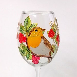 Puede incluir: Una copa de vino transparente con un diseño pintado a mano que presenta un petirrojo posado en una rama con frambuesas rojas y hojas verdes. El pájaro tiene el pecho naranja y el cuerpo gris. La copa es de cristal.